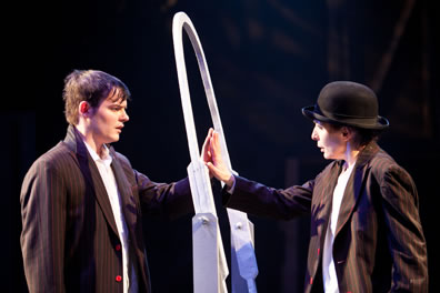 Sebastian and Viola touch palms through a white oval mirror frame, both in pinstripe jackets and white shirts, she in bowler hat.